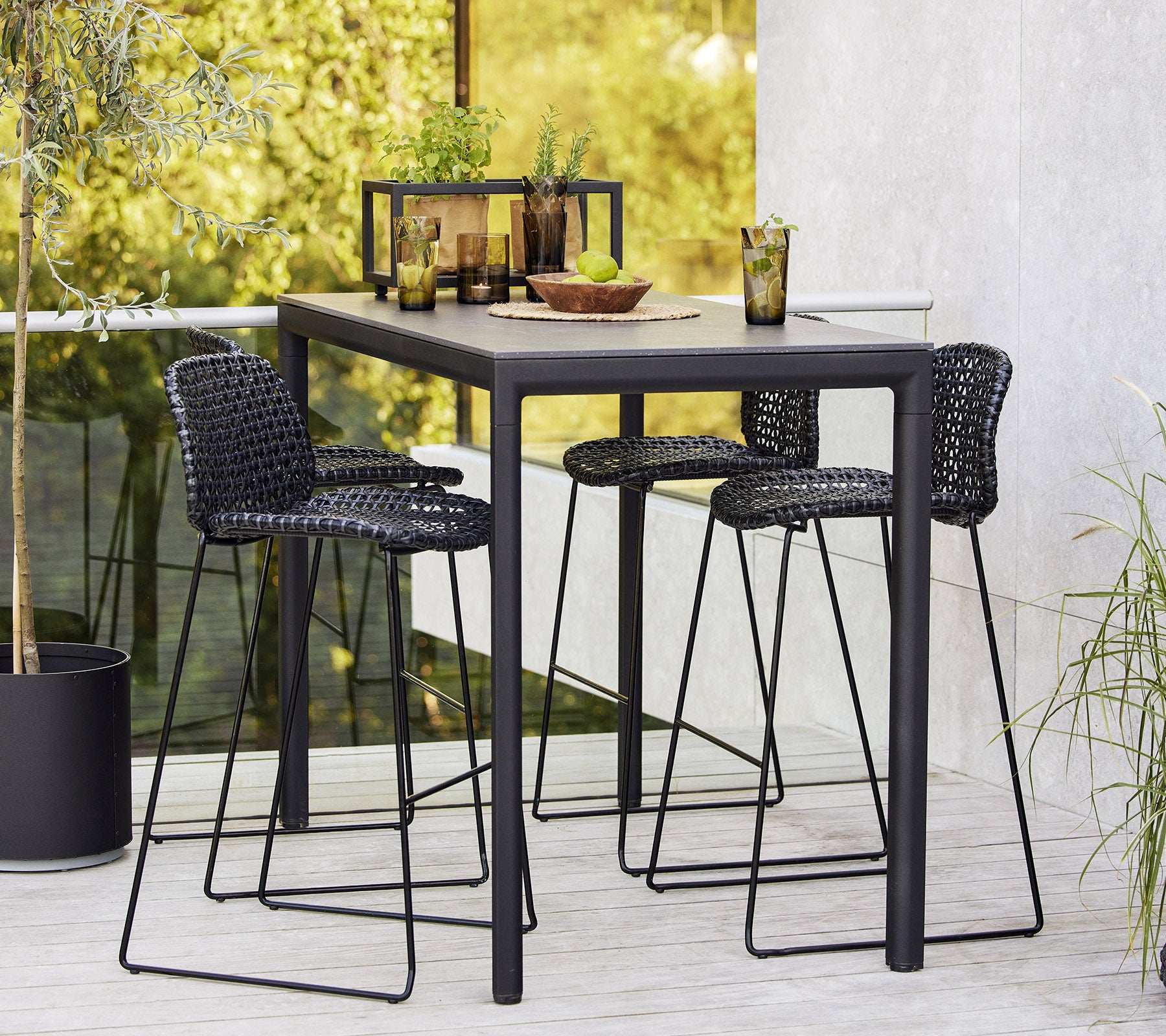 A stylish drop bar table set with three woven bar stools, arranged with tableware and drinks, surrounded by greenery.