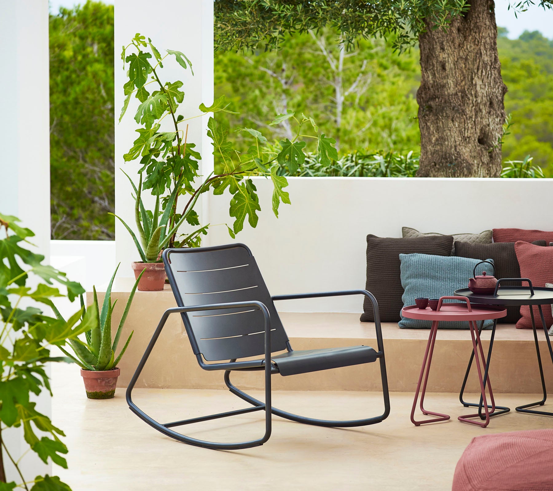 Stylish rocking chair beside potted plants and cushions, creating a cozy and inviting seating area with a serene view.