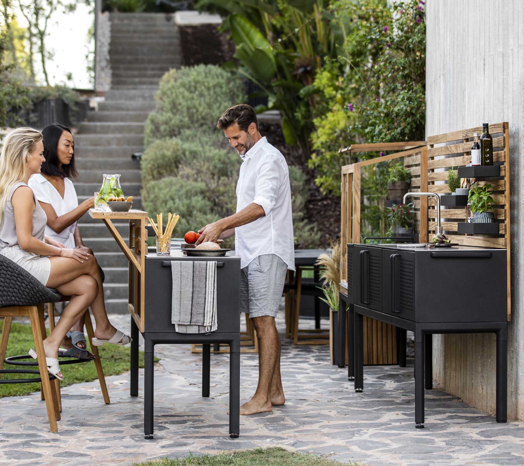 A man prepares food at a modern outdoor kitchen, while two women enjoy drinks nearby, surrounded by greenery and stylish decor.