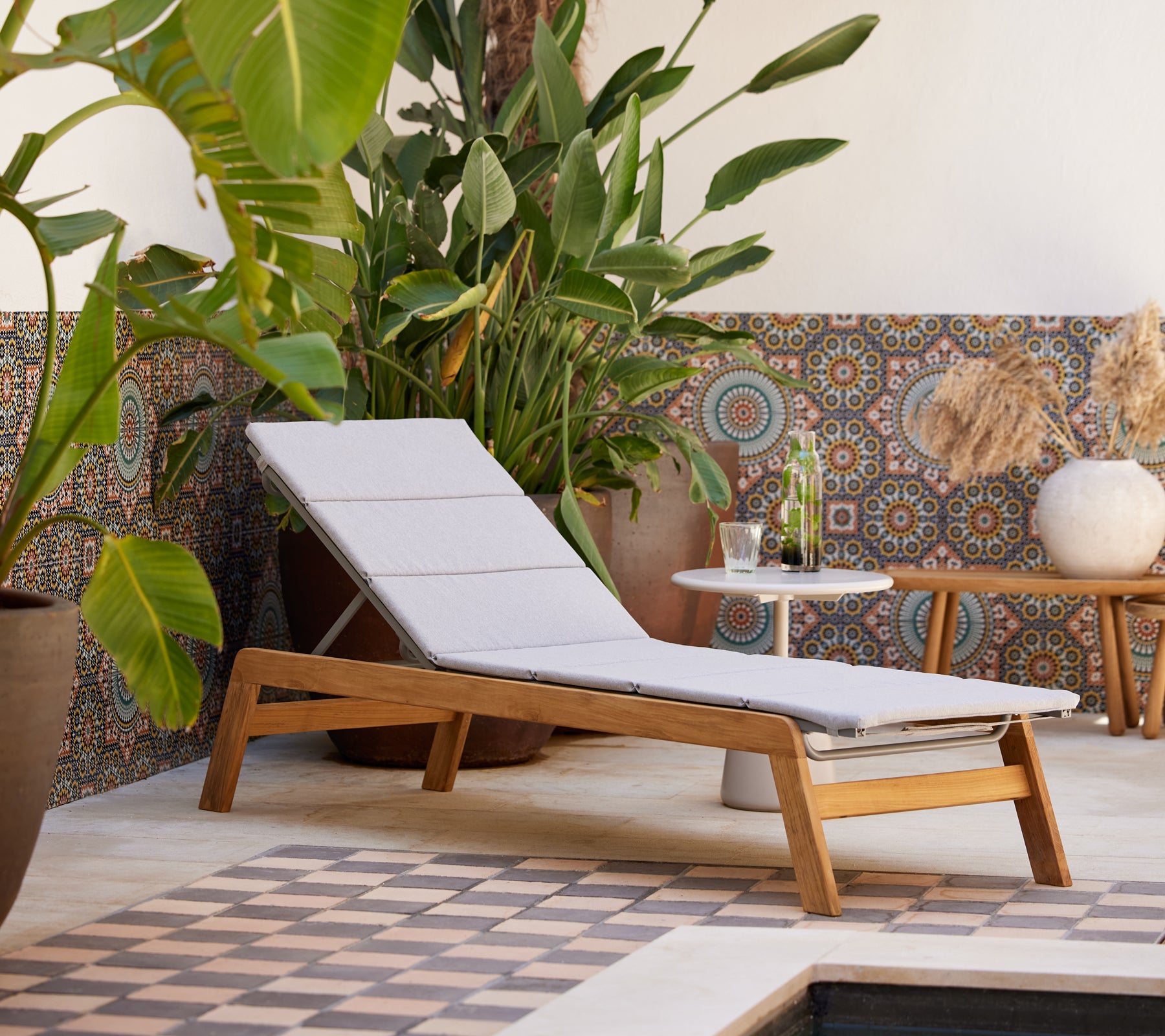 A modern lounge chair with a light cushion, paired with a side table, surrounded by lush greenery and decorative tiled background.