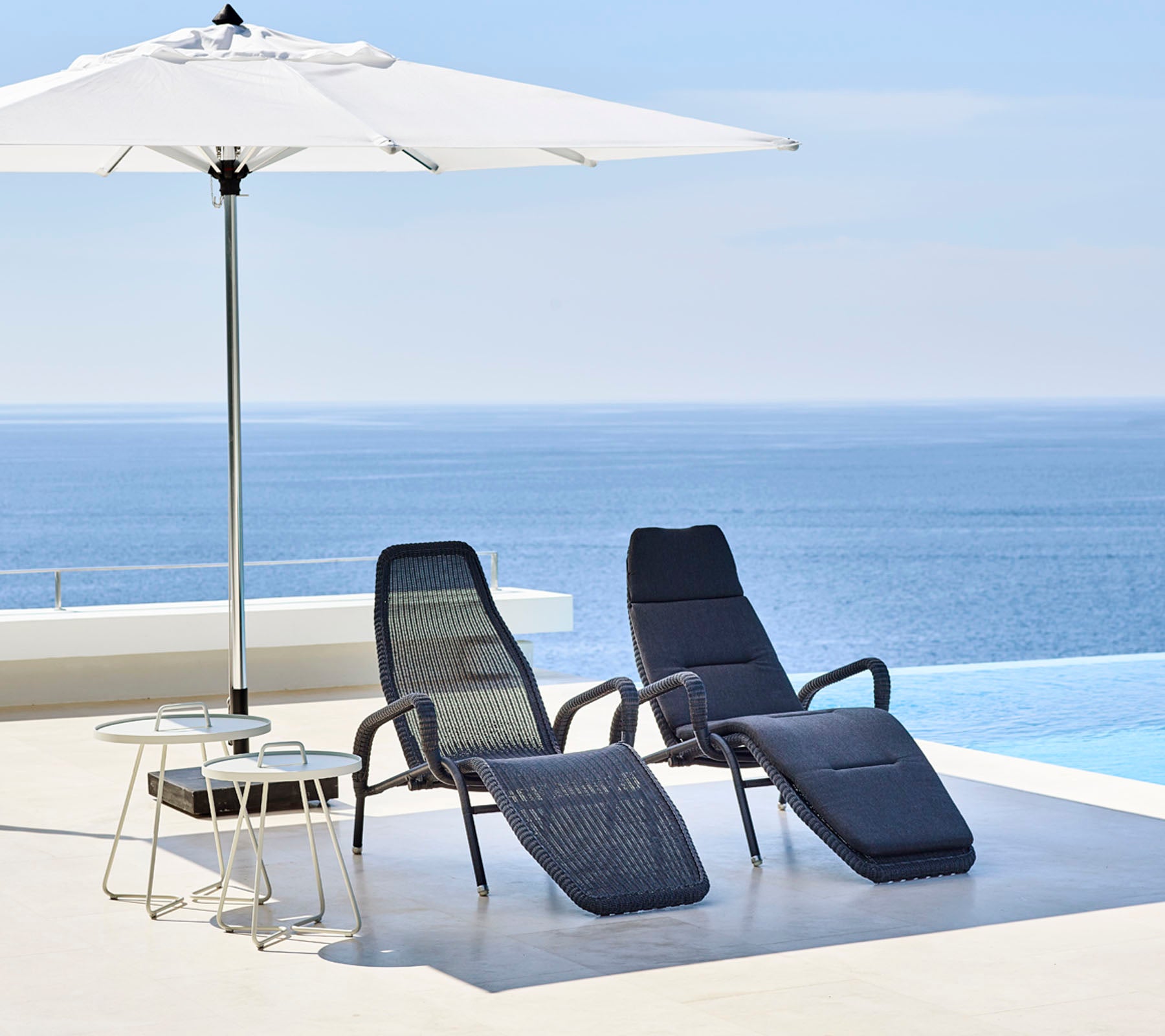 Two stylish lounge chairs and a small table sit under a large umbrella, with a serene blue sea in the background.
