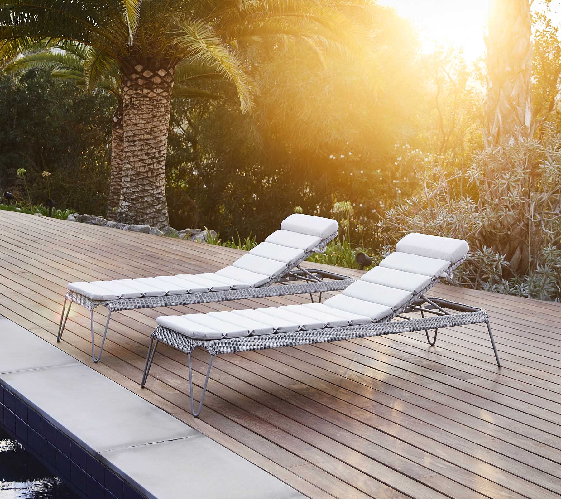 Two stylish lounge chairs with white cushions, positioned beside a pool and surrounded by lush greenery, basking in warm sunlight.