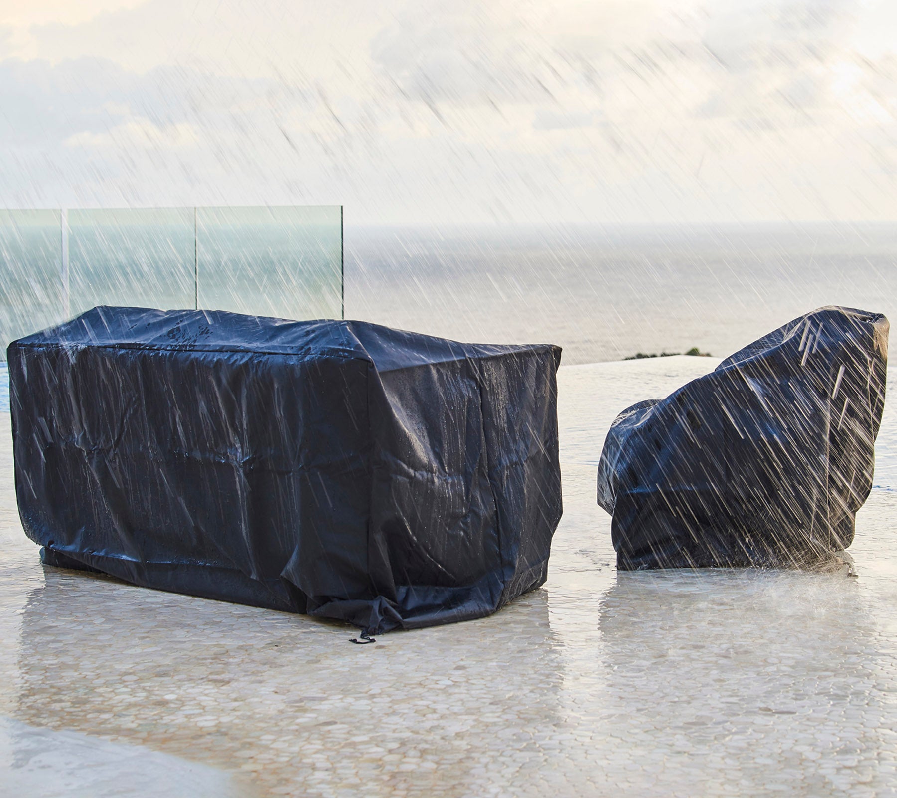 Outdoor furniture covered with dark protective covers, shielded from rain, with a backdrop of a cloudy sky and ocean.