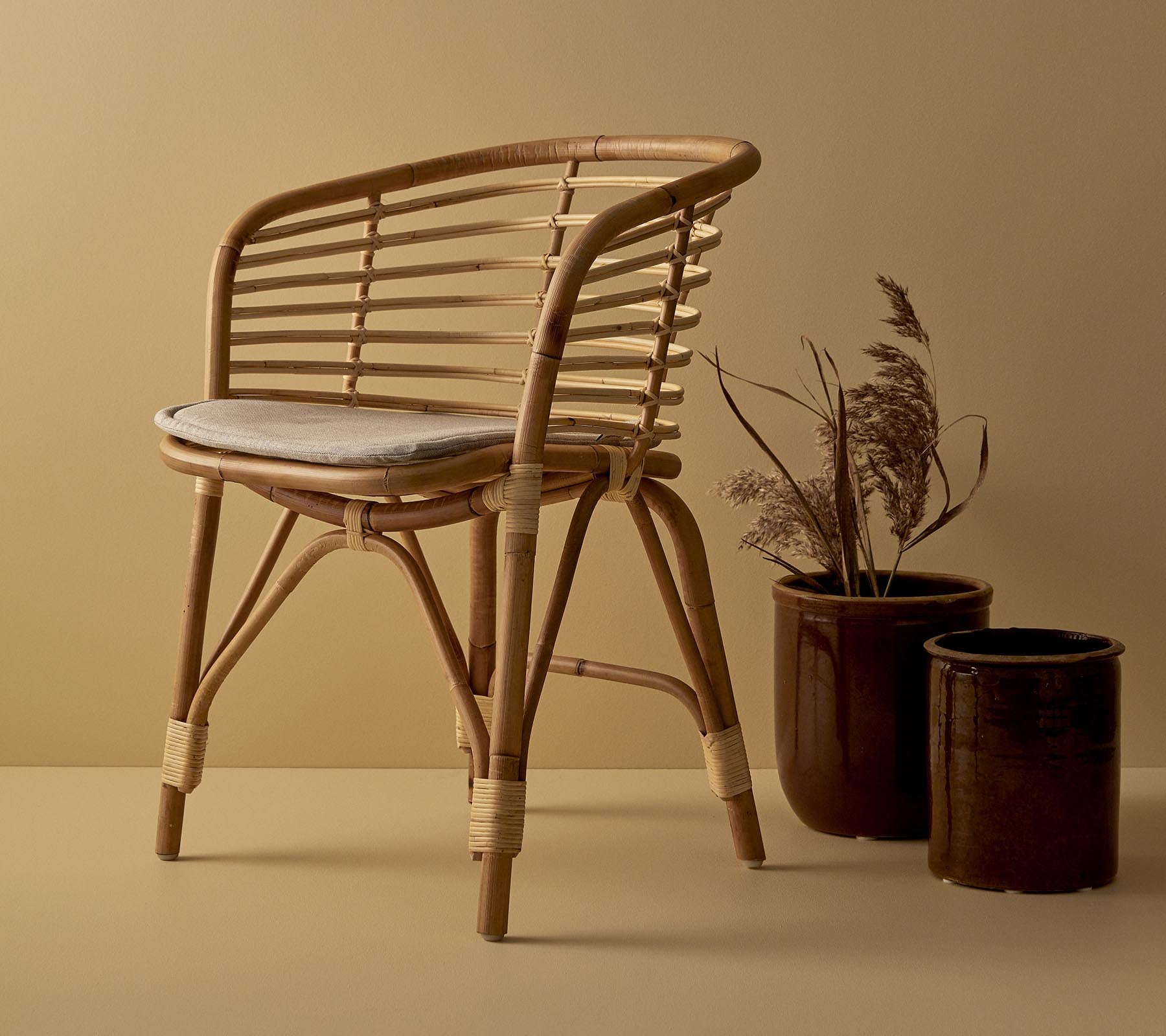 A stylish rattan chair with a cushioned seat, accompanied by two decorative planters featuring dried plants.