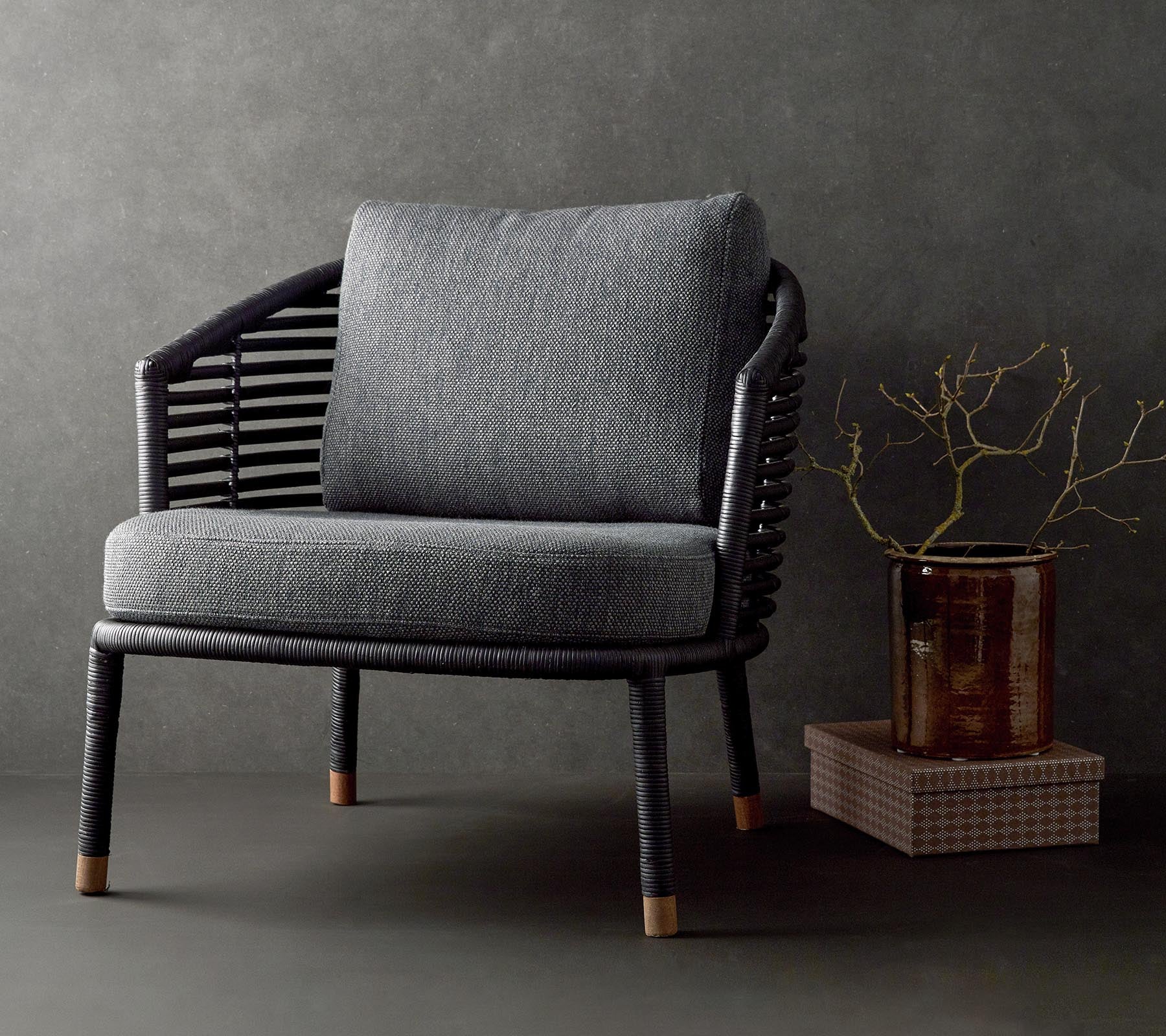 Stylish chair with a textured gray cushion and woven black frame, complemented by a decorative branch and a small table.