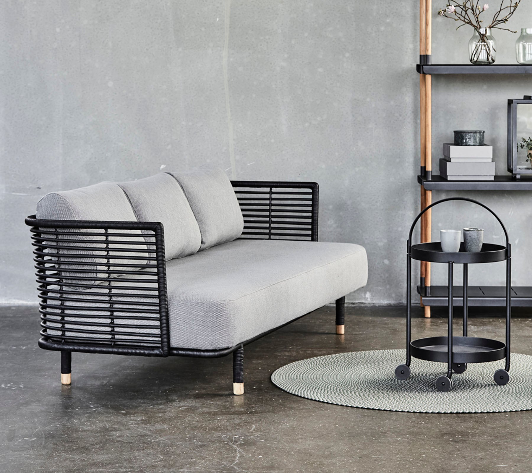 Sleek gray sofa with black metal accents and cushions, paired with a round side table on a textured rug against a neutral background.