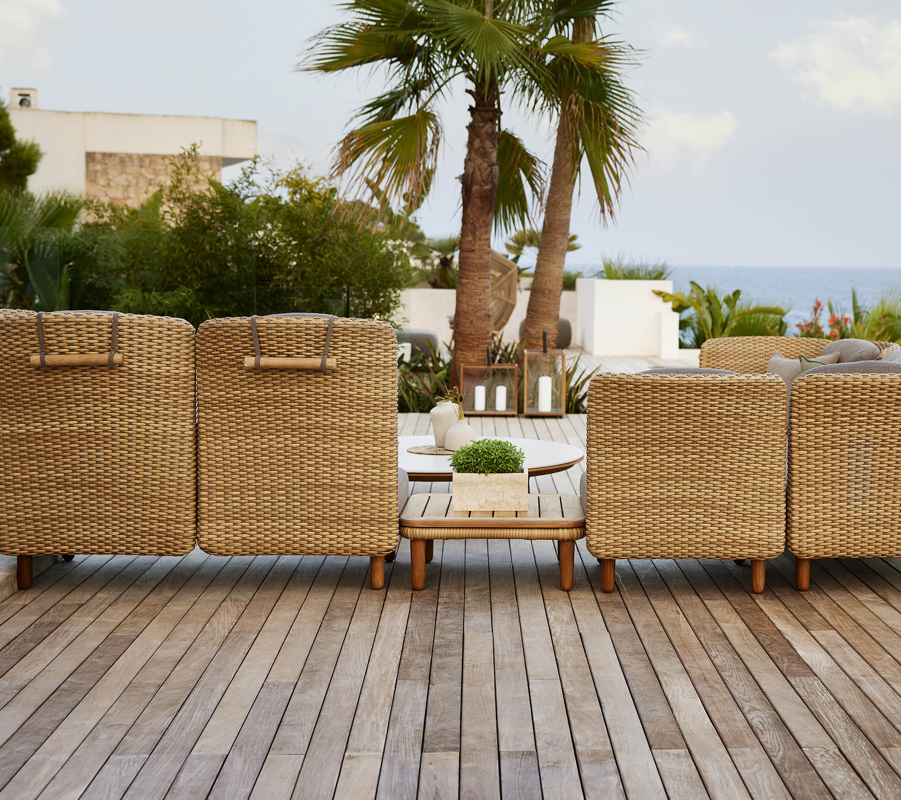 A row of woven lounge chairs facing a scenic view, with a wooden deck and tropical plants in the background.