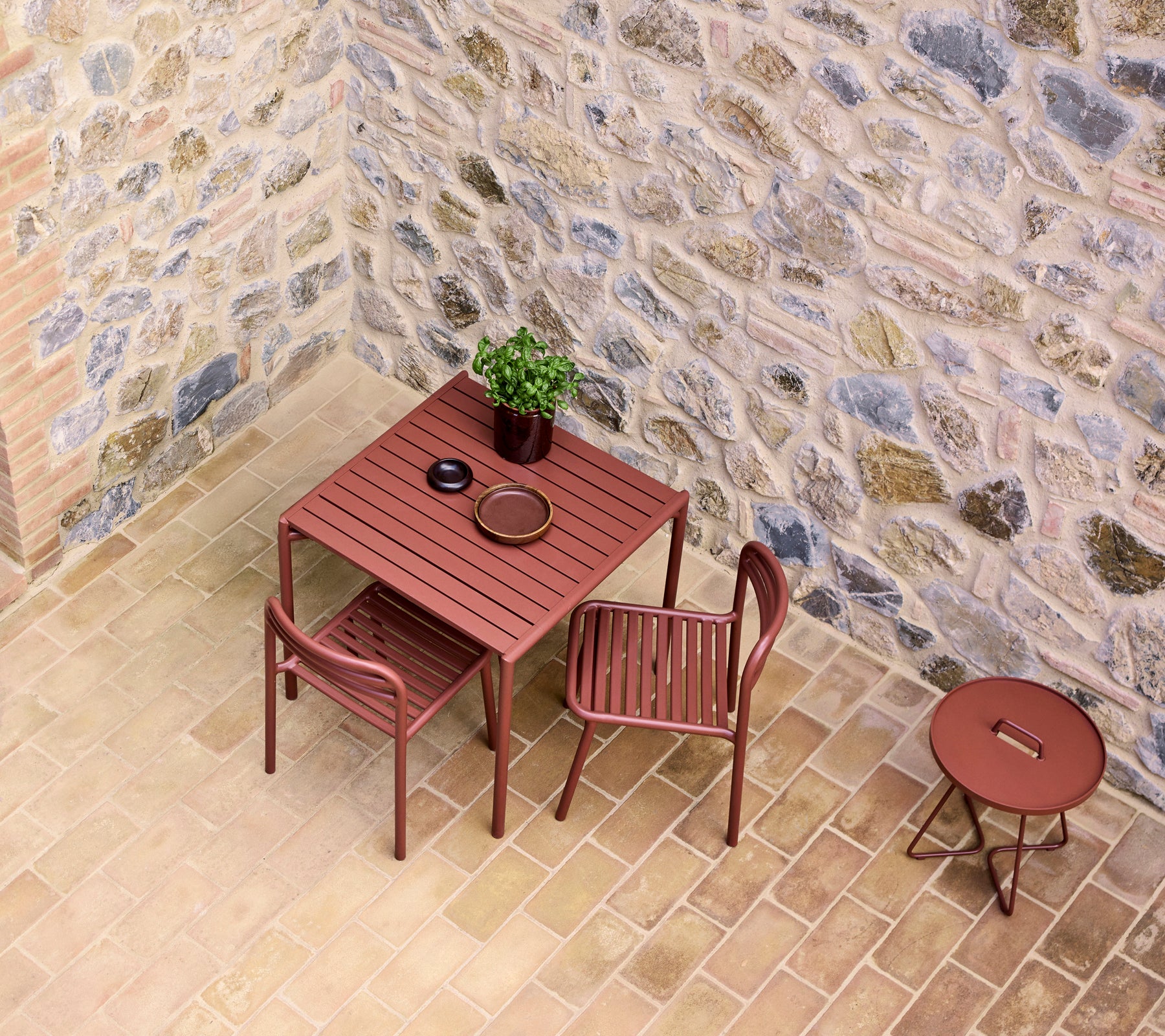 A red cafe table set with two chairs, featuring a small plant and a plate, against a stone wall backdrop.