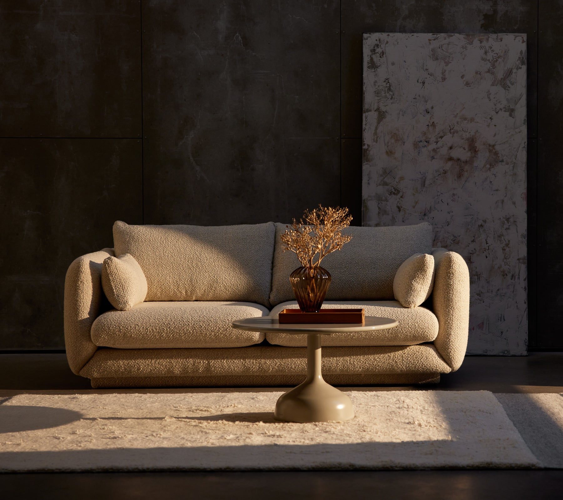 Bloom 2-seater sofa in sand flair with a Glaze coffee table and decorative plant on a rug, set against a textured dark wall.