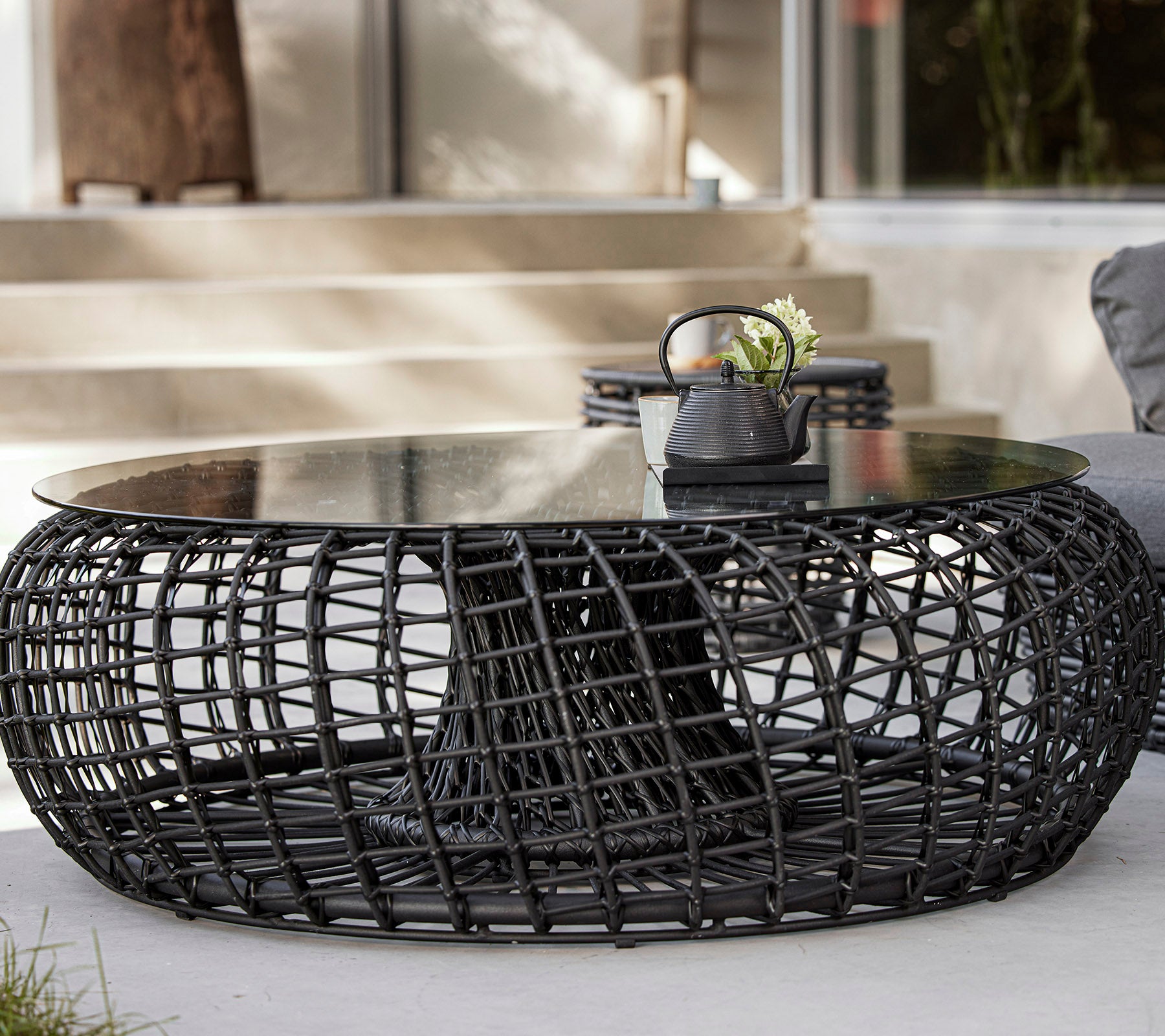 A modern, round coffee table with a woven black base and a glass top, featuring decorative items like a plant and a small bowl.