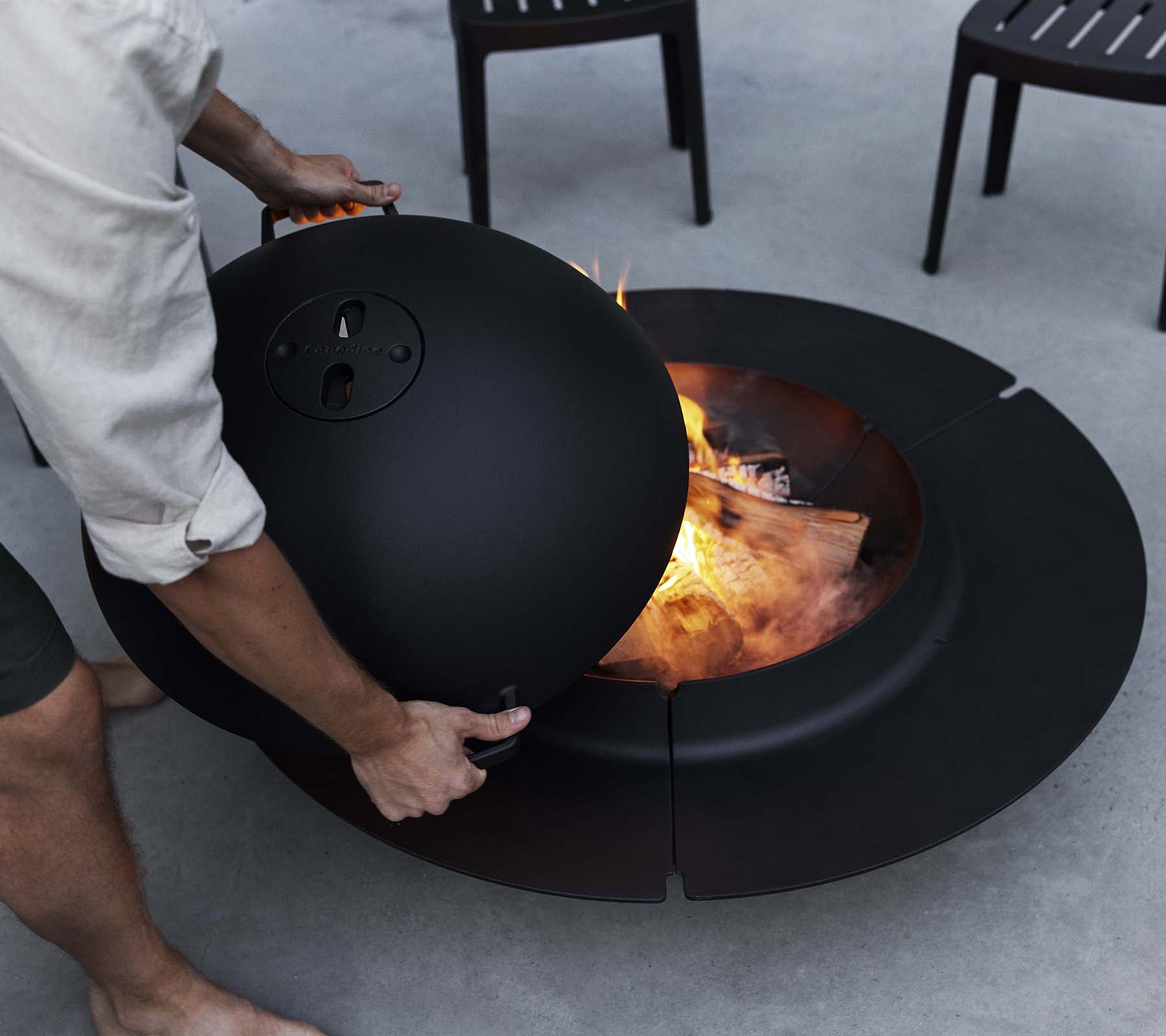 A person adjusts a lid on a sleek black Ember fire pit with flames and burning wood visible inside, surrounded by chairs.