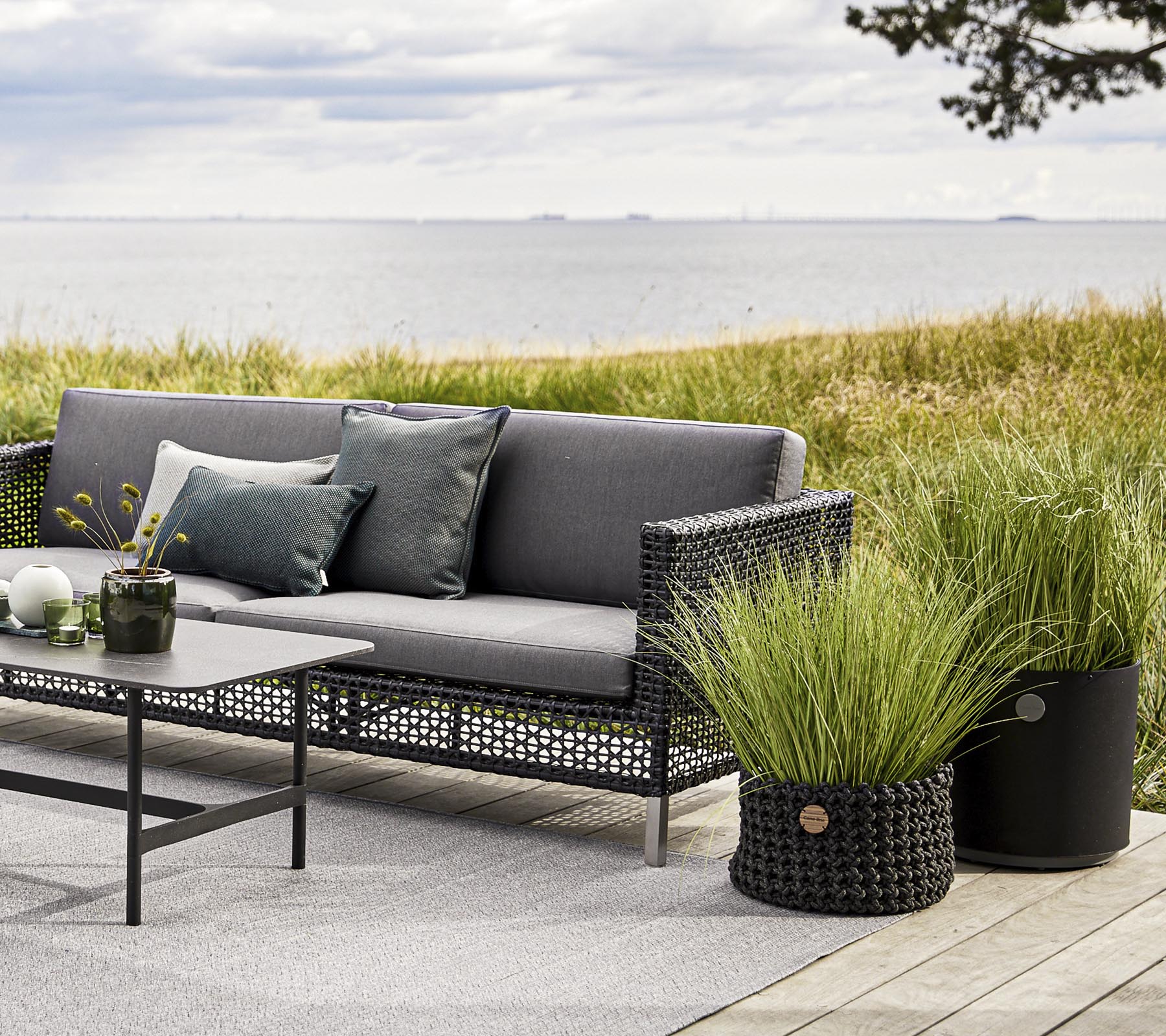 Stylish outdoor seating with a gray couch, decorative pillows, and a table, complemented by decorative grass planters and a serene backdrop.