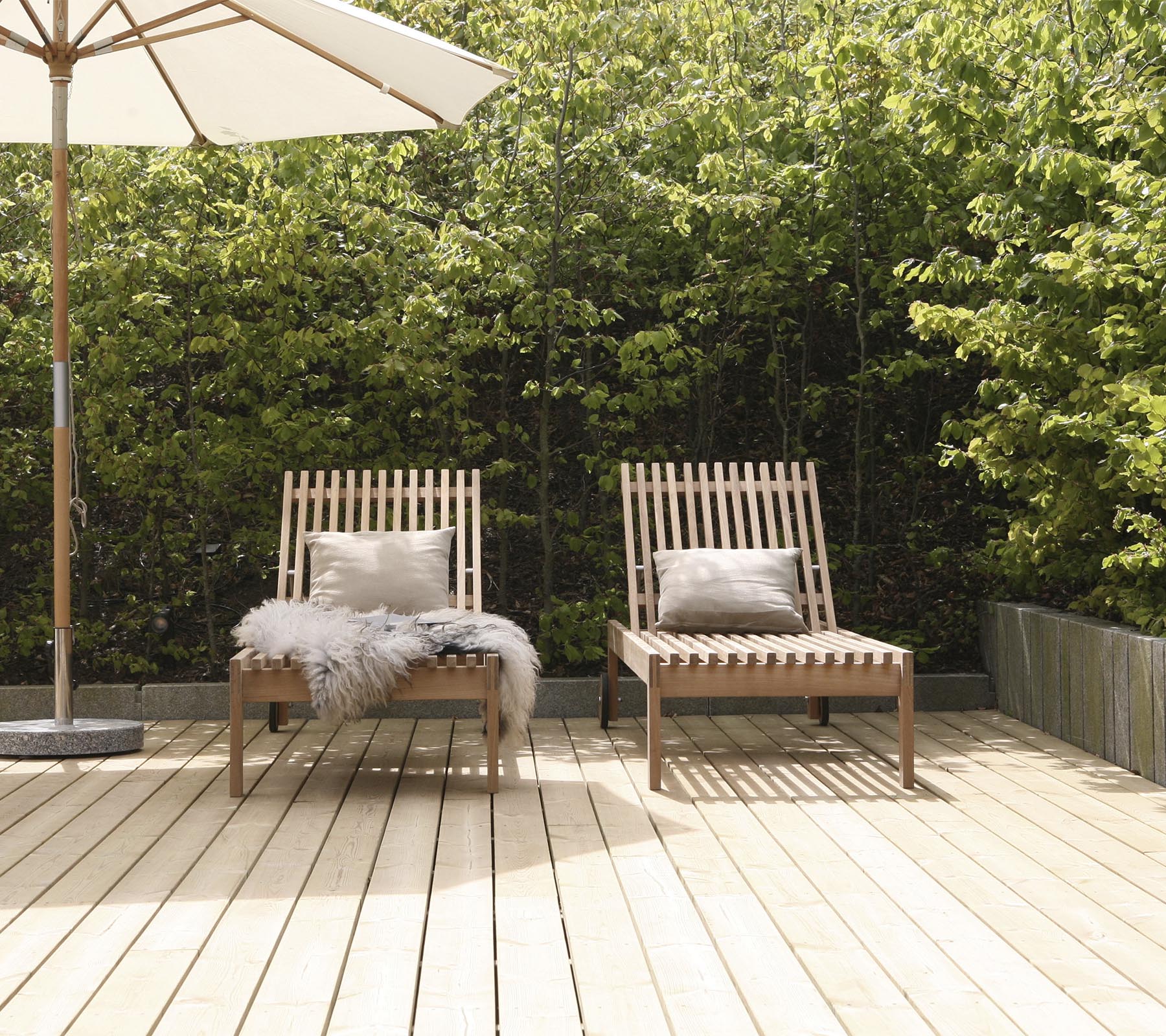 Two stylish wooden lounge chairs with cushions, positioned on a wooden deck, surrounded by greenery and a large umbrella nearby.