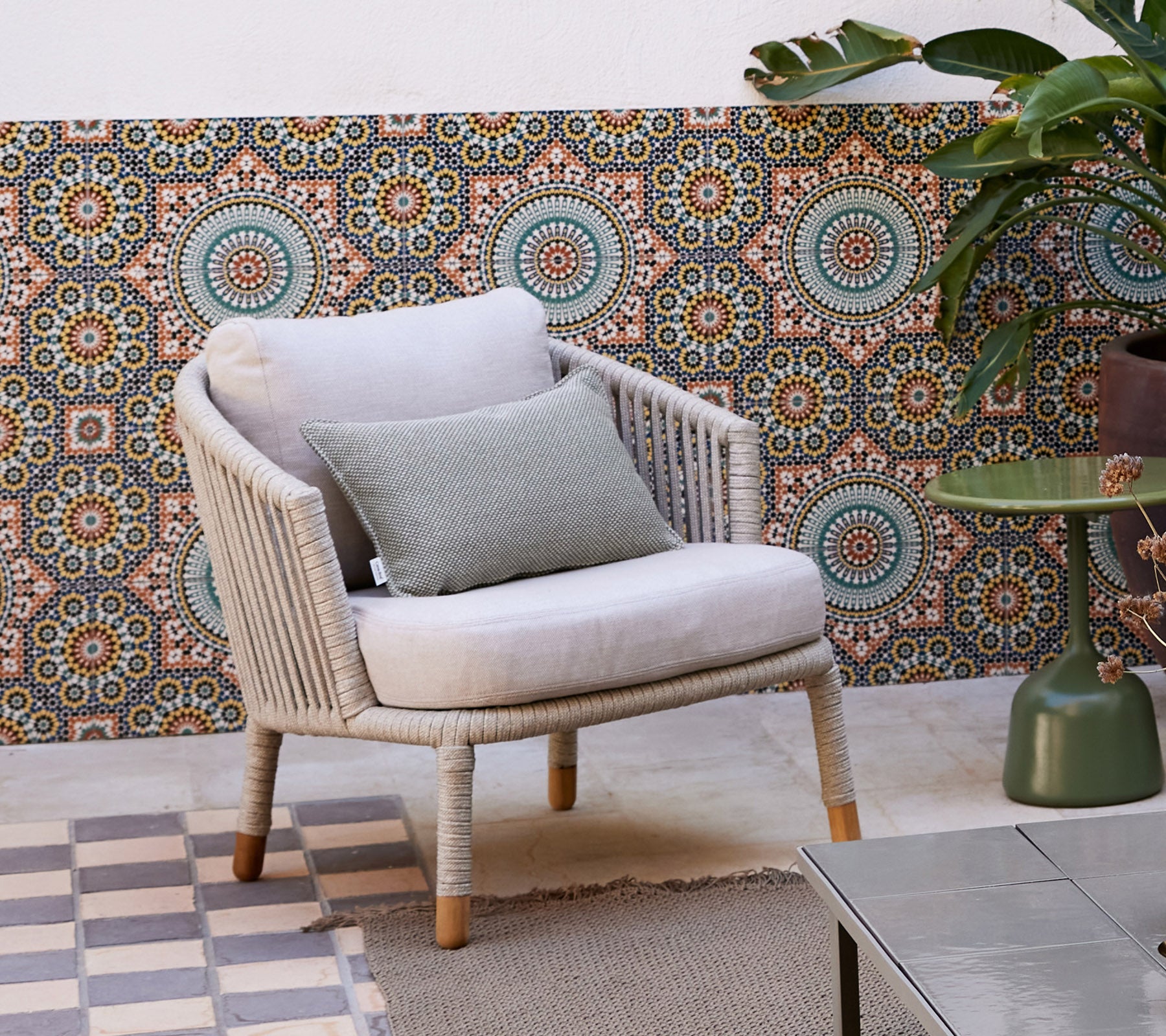 A stylish lounge chair with a soft cushion and a decorative pillow, set against a patterned backdrop and surrounded by a potted plant.